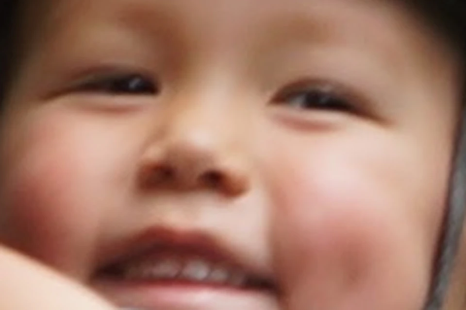 A smiling child in a helmet and harness glides on a zip line through a forest; another person waits on a platform in the background.