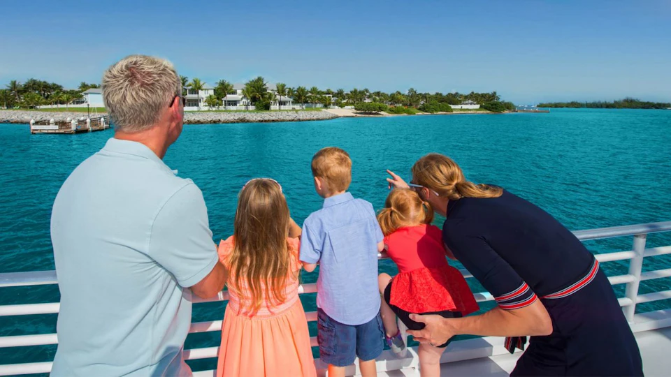 A family of four looks across the water towards a distant shoreline with palm trees and buildings. One child points towards the shore.