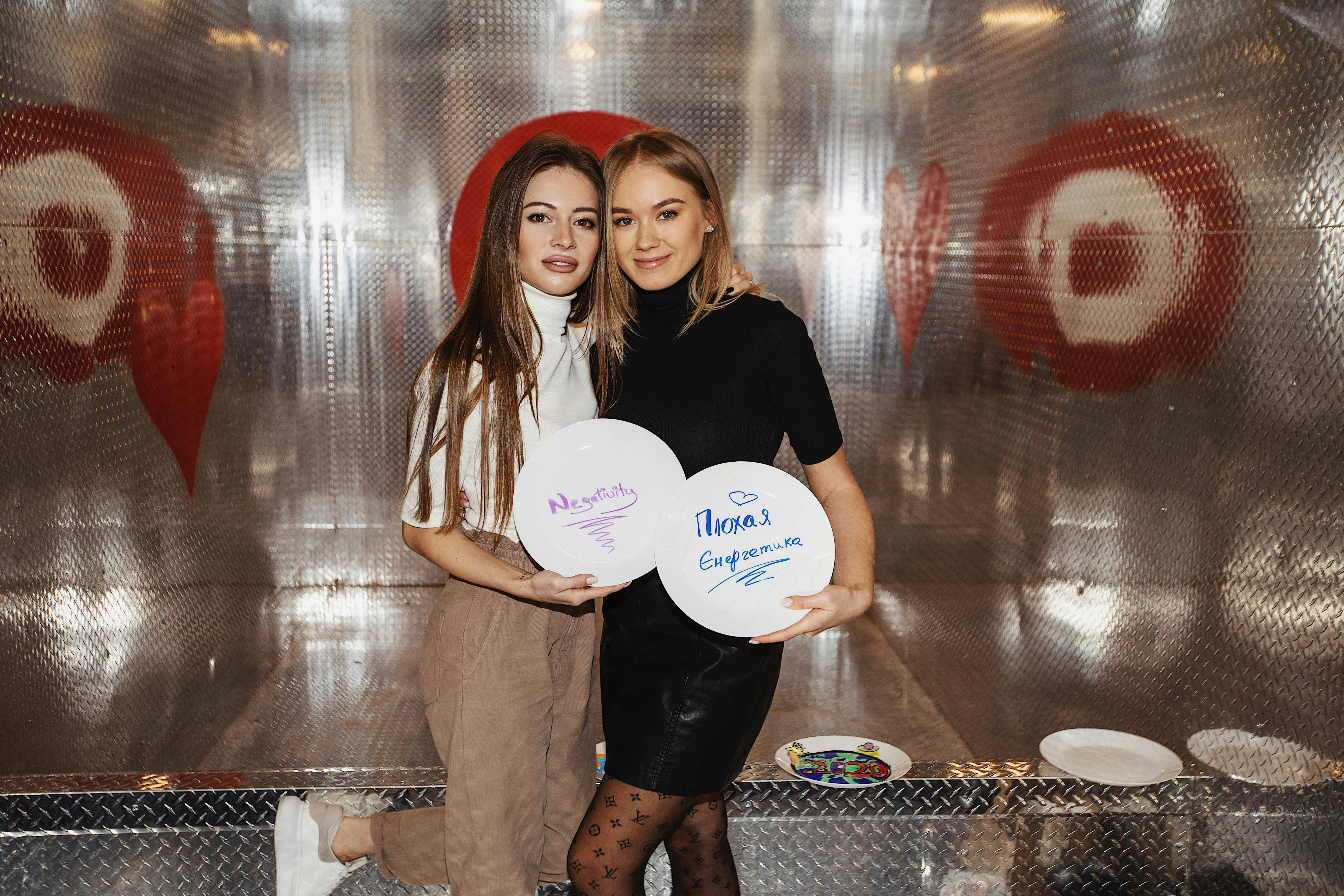 Dues dones joves posen juntes sostenint plats blancs amb missatges escrits a mà. El fons presenta parets metàl·liques amb dissenys de cor.