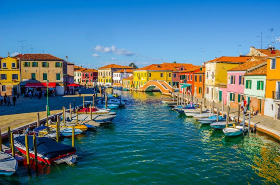A canal lined with colorful buildings and boats, with a small arched bridge in the center under a clear blue sky.