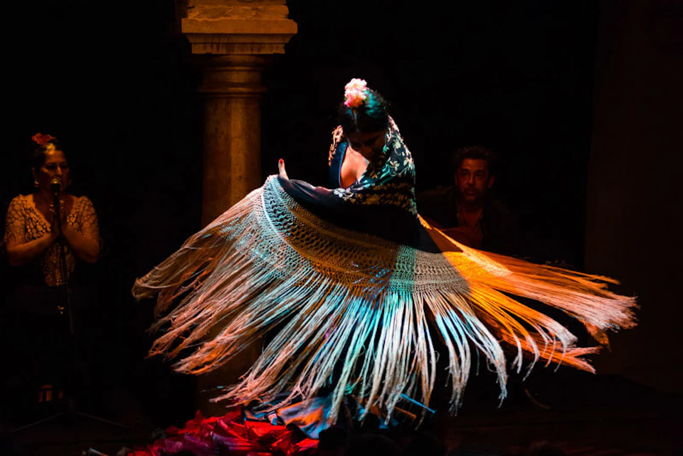 A dancer with a flower in her hair gracefully twirls a shawl with long fringes on a dimly lit stage, with a man in the background.