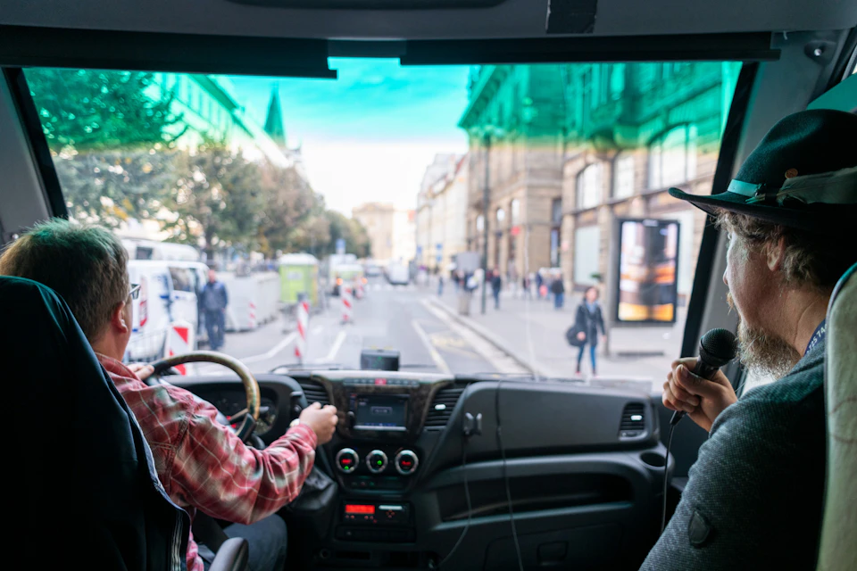 View from inside a vehicle driving through a city street with pedestrians and buildings on either side.