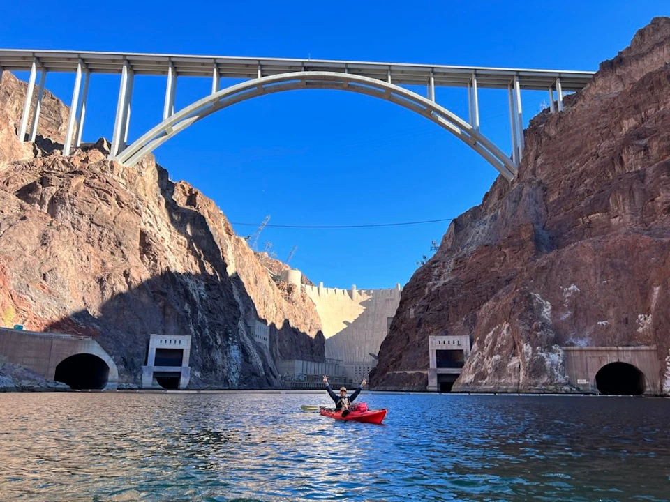 A person in a red kayak paddling on water beneath a large arched bridge, with cliffs and a dam in the background.