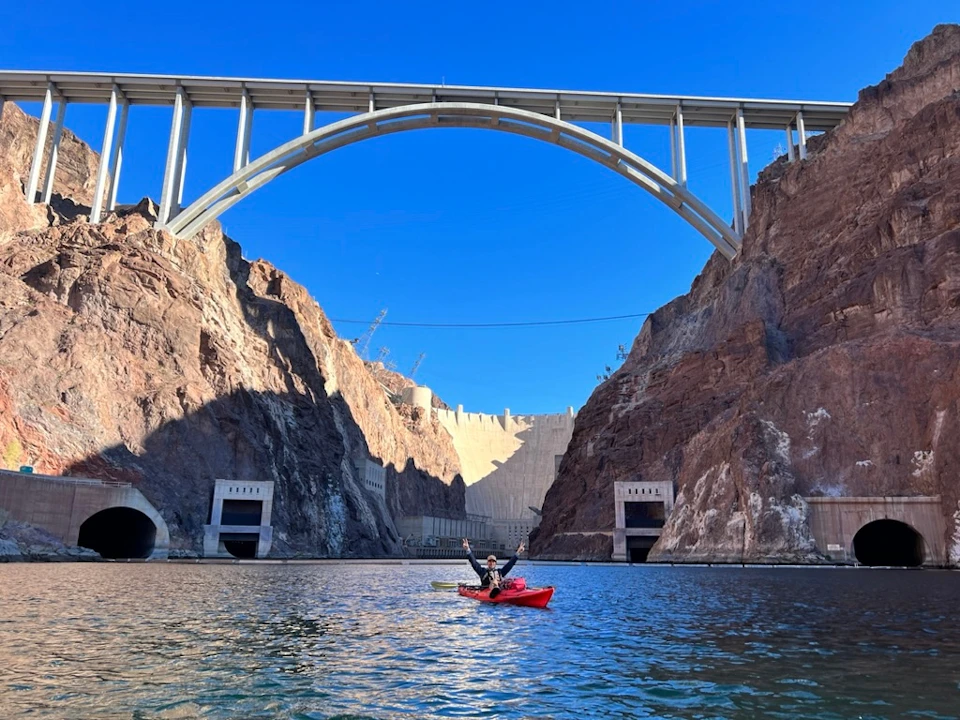 A person in a red kayak paddling on water beneath a large arched bridge, with cliffs and a dam in the background.