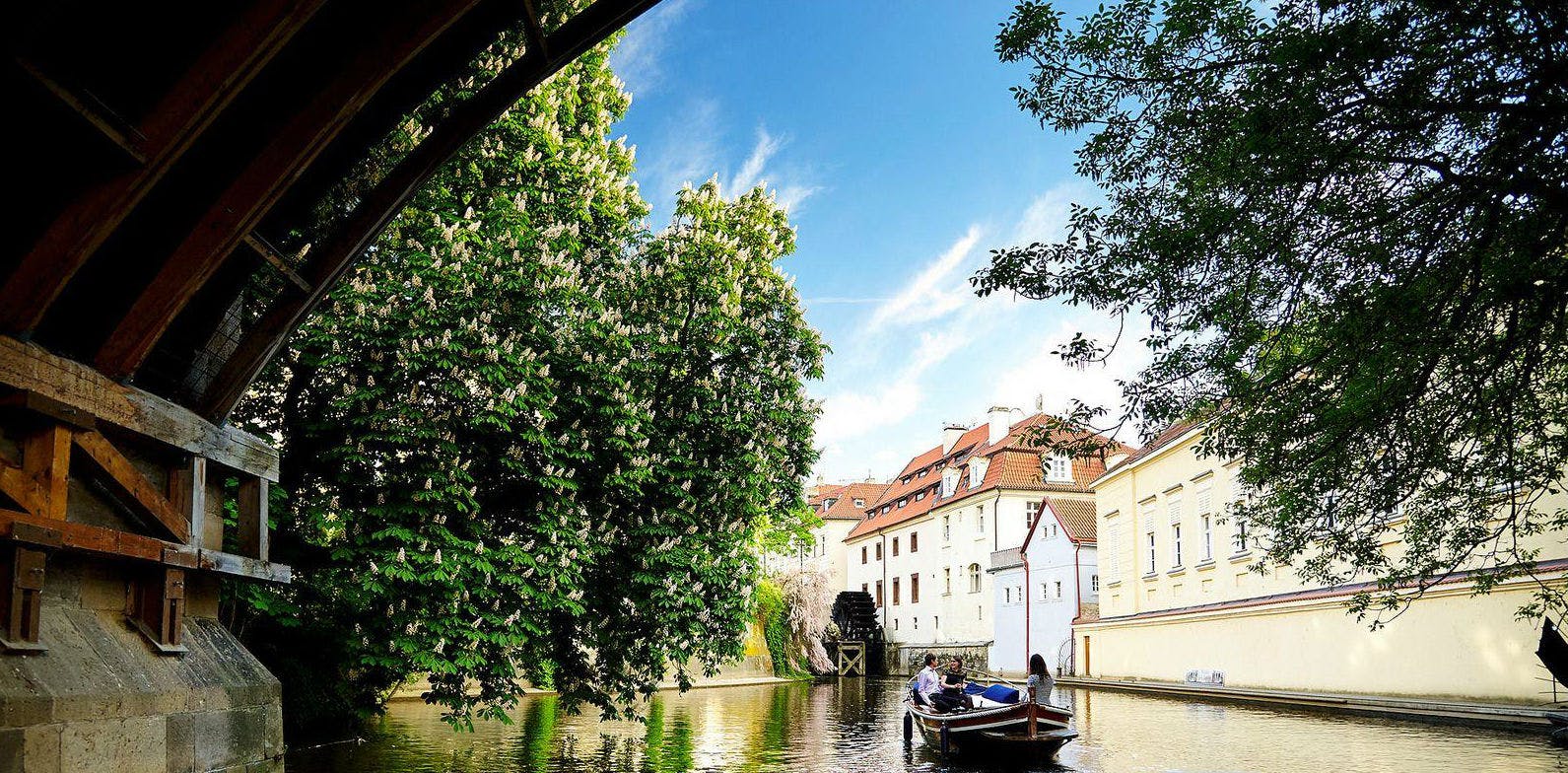 A small boat with people on a calm river, surrounded by lush trees and historic buildings under a clear blue sky.