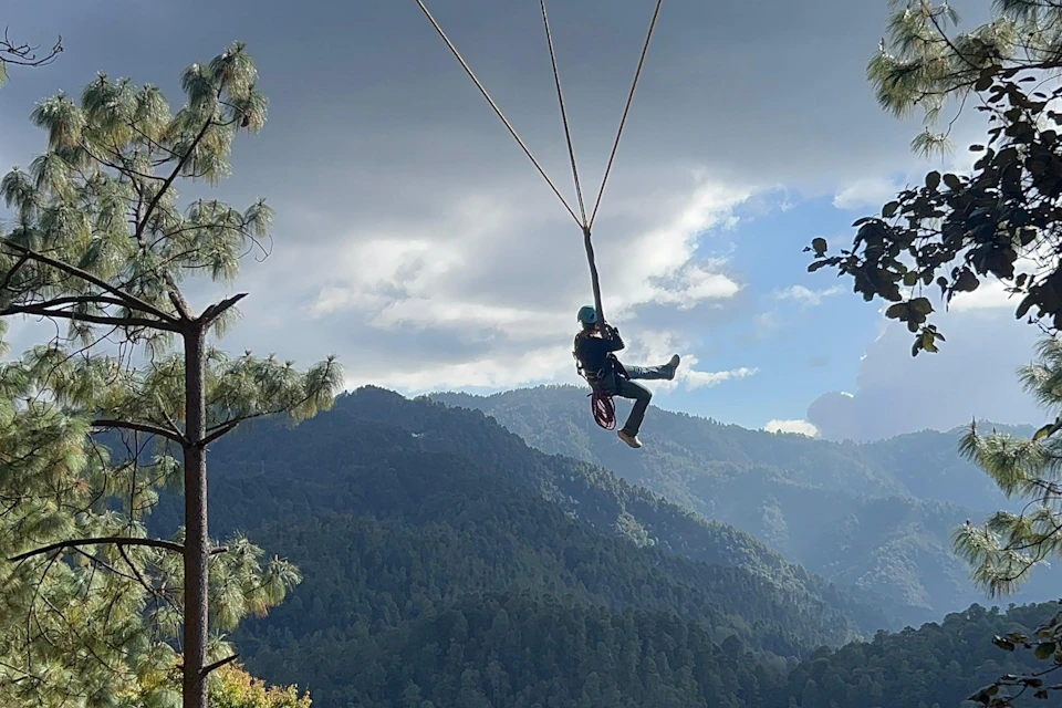 A person suspended on a zipline set against a forested mountain landscape under a partly cloudy sky.