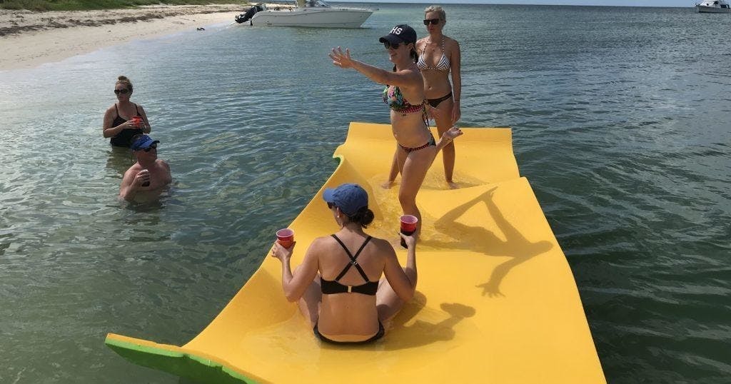 Três mulheres num tapete flutuante amarelo em águas pouco profundas perto de uma praia, com um barco visível ao fundo.