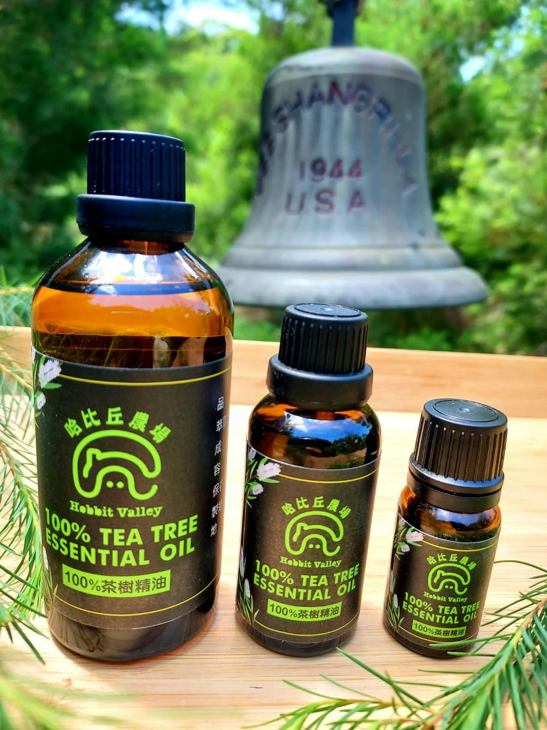 Three bottles of Hobbit Valley 100% Tea Tree Essential Oil on a wooden surface, with a blurred outdoor background.