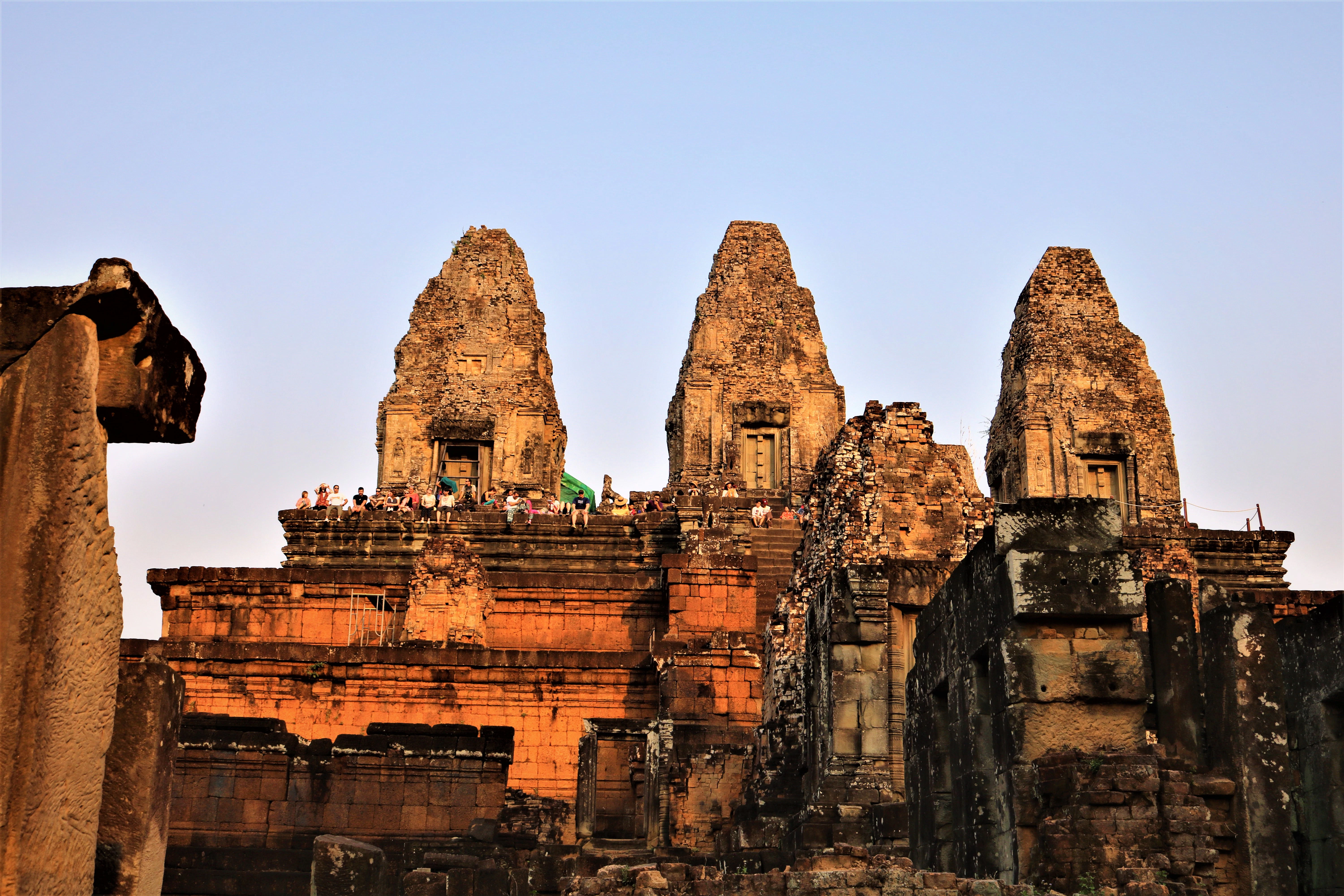 Ancient temple ruins with three tall stone towers at sunset, and a few people standing near the top. Clear, blue sky above.
