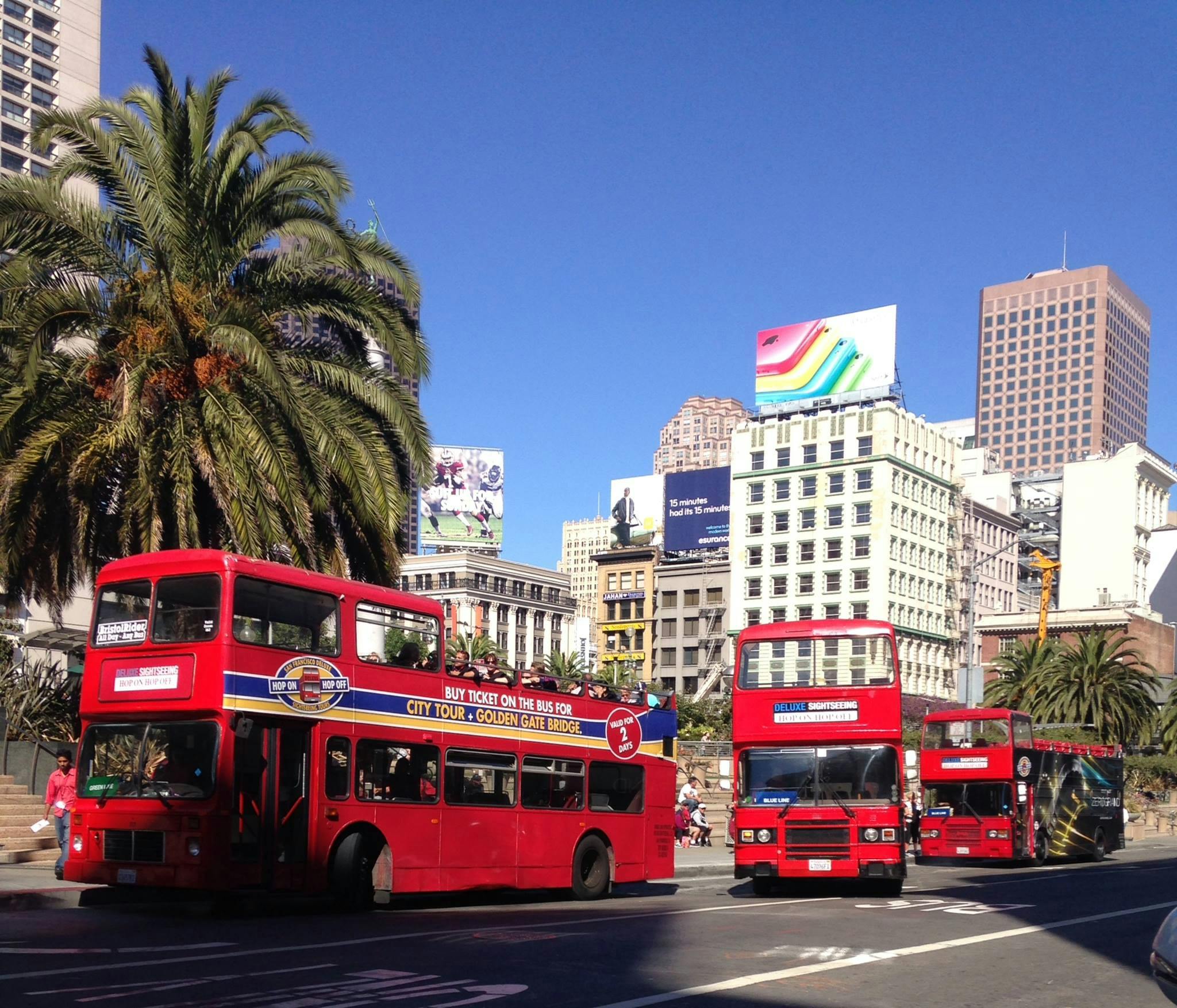 Plusieurs bus rouges à impériale circulent dans une rue de la ville bordée de palmiers et de bâtiments modernes, sous un ciel bleu clair.