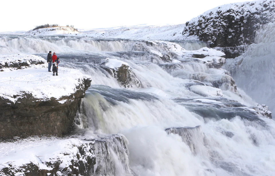 A snowy landscape with a cascading waterfall and two people in winter clothing standing on a cliff edge.