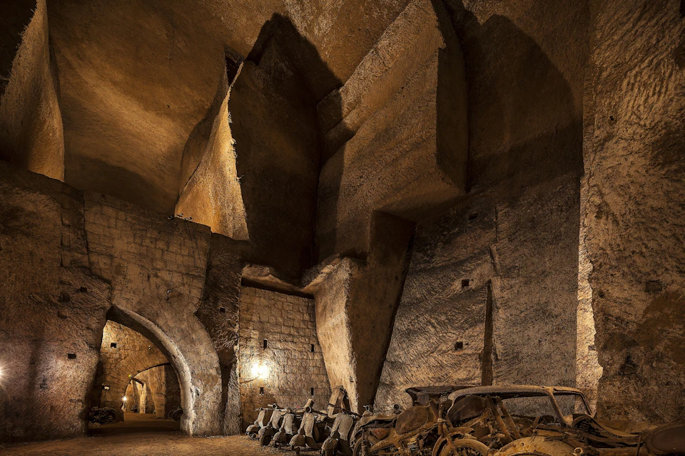 An underground cavern with stone walls and ceiling, lit by wall-mounted lights, with a row of old motorcycles covered in dust.