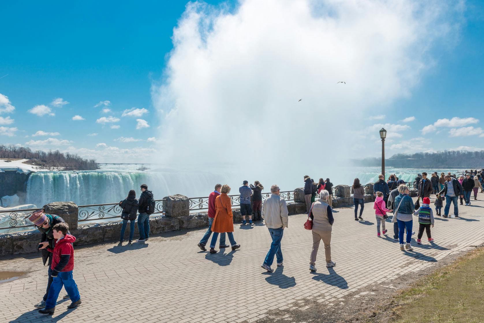 Personnes se promenant et observant les chutes du Niagara depuis une aire d'observation par une journée ensoleillée, la brume s'élevant de la chute d'eau à l'arrière-plan.
