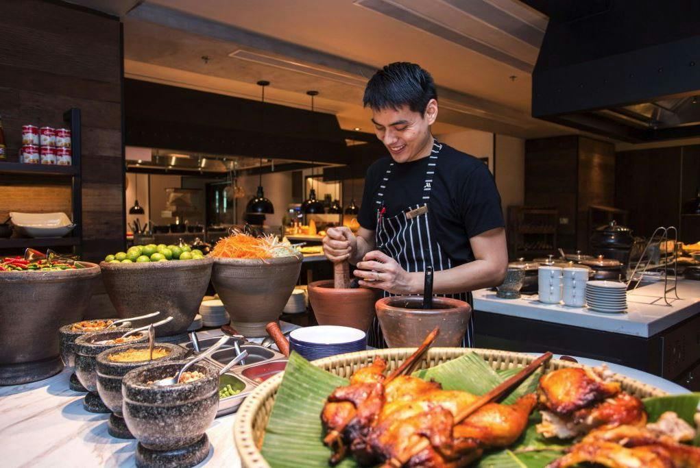 A person wearing a striped apron is using a mortar and pestle at a food station with various dishes, including grilled skewers.