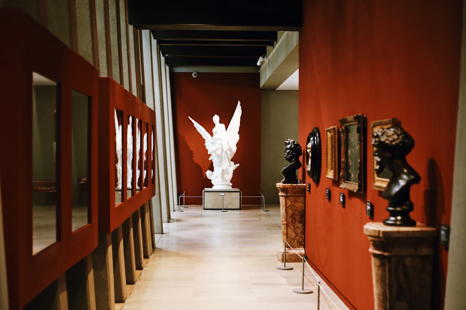 A museum hallway with a white winged statue at the end, red walls, mirrors on the left, and various sculptures and framed art on the right.