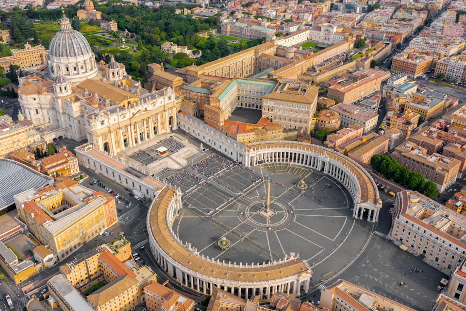 Aerial view of St. Peter's Square in Vatican City, showing the basilica, colonnades, central obelisk, and surrounding buildings.