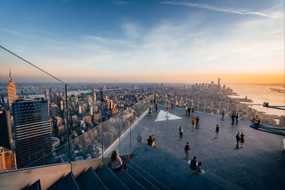 People on a rooftop observation deck overlooking a city skyline at sunset, with glass railings and wide concrete steps.