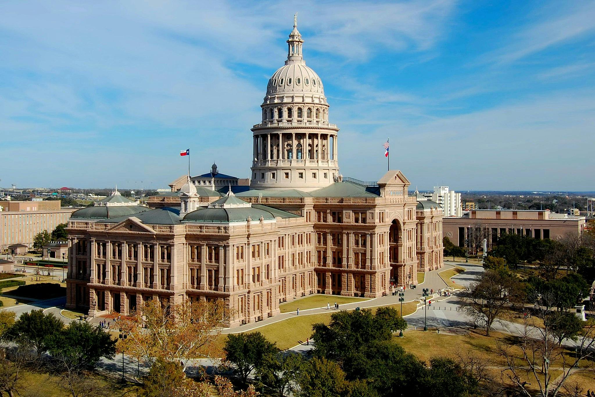 The image shows a large, historic building with a prominent dome and multiple flags, surrounded by lawn and trees under a blue sky.