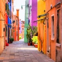 Narrow alleyway with colorful buildings, potted plants, and a tiled pathway illuminated by natural daylight.