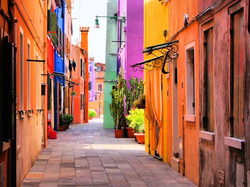 Narrow alleyway with colorful buildings, potted plants, and a tiled pathway illuminated by natural daylight.