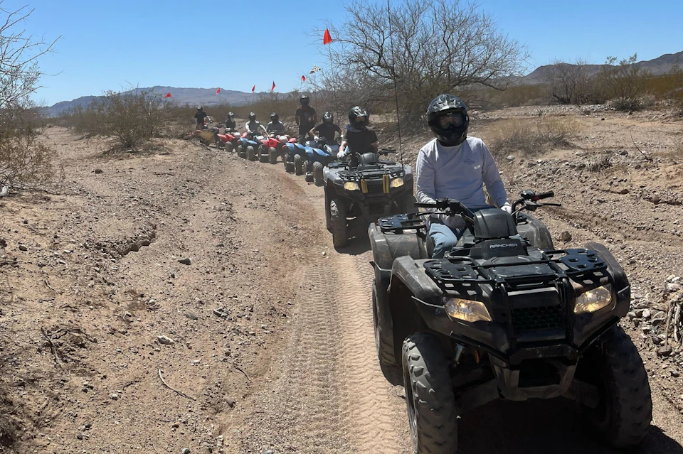 A group of people riding all-terrain vehicles on a dirt trail in a desert landscape under a clear blue sky.
