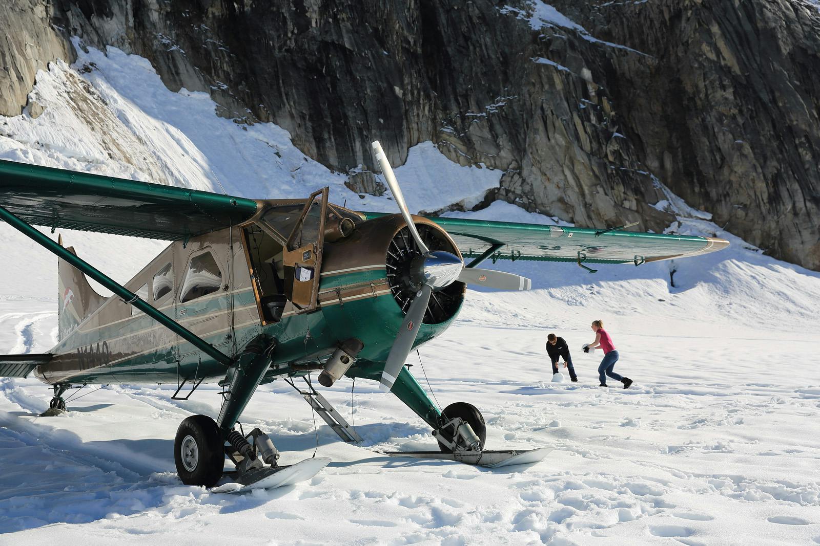 Un avion de brousse vert avec des skis est garé sur la neige près d'une montagne rocheuse ; deux personnes marchent à proximité.