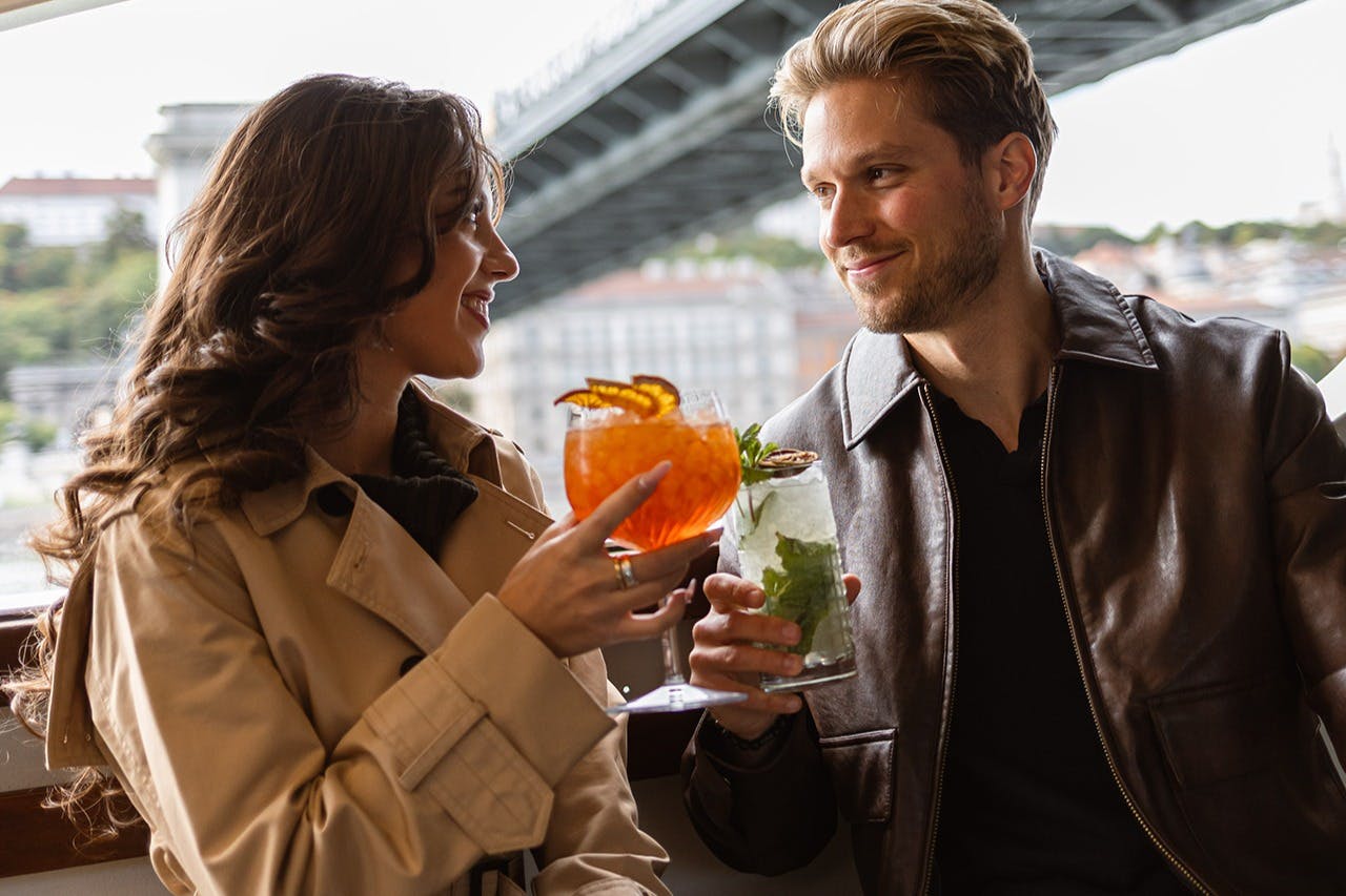 A man and woman smile at each other while holding cocktails, with a bridge visible in the background.