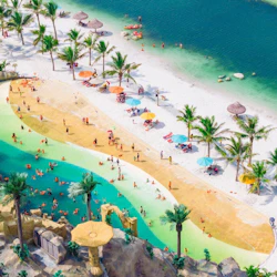 Aerial view of a beach with people swimming, sunbathing, and relaxing under umbrellas and palm trees by turquoise water.