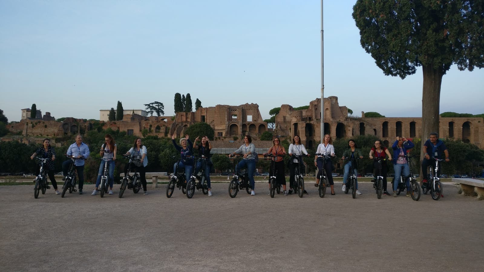 Un gruppo di persone in bicicletta posa per una foto di fronte ad antiche rovine e alberi sotto un cielo limpido.