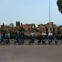 Un gruppo di persone in bicicletta posa per una foto di fronte ad antiche rovine e alberi sotto un cielo limpido.