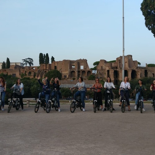 Un gruppo di persone in bicicletta posa per una foto di fronte ad antiche rovine e alberi sotto un cielo limpido.