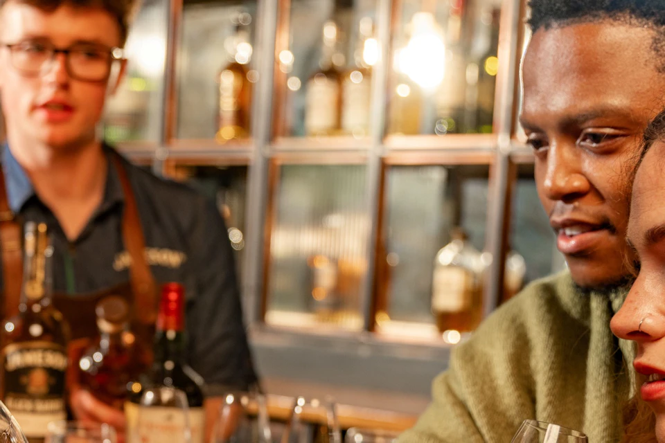 People tasting whiskey at a bar with a bartender in the background, surrounded by bottles displayed on shelves.