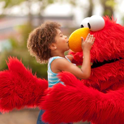 A child in a blue dress kisses a person in an Elmo costume outdoors, with greenery in the background.
