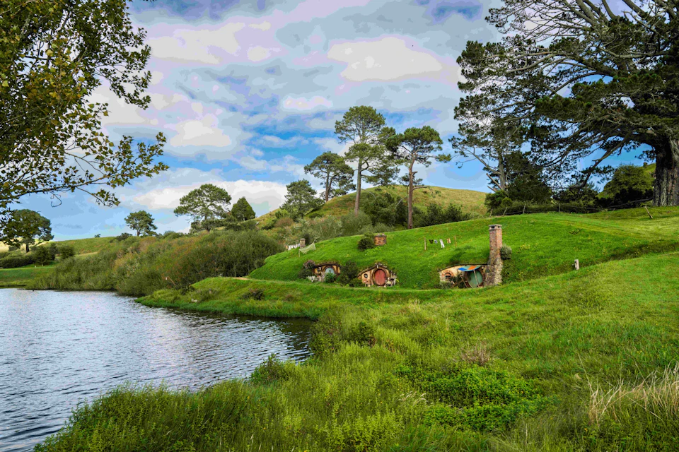 A scenic hillside with small round-door houses, a pond, trees, and laundry hanging outdoors beneath a cloudy sky.