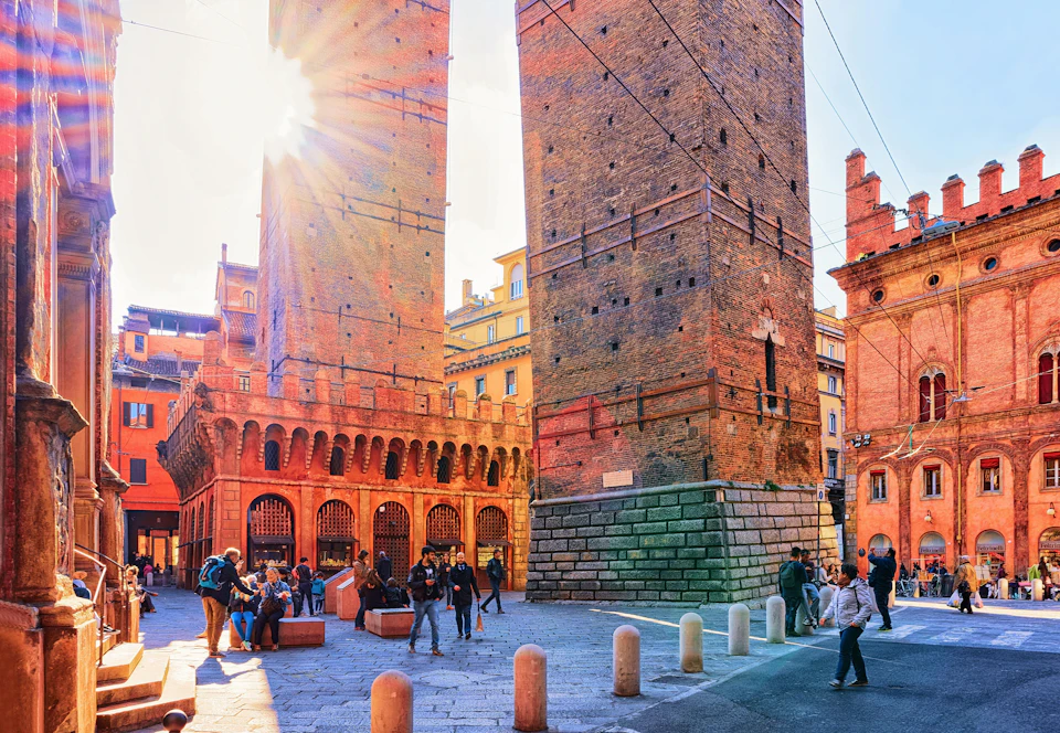 People walking and gathering near historic twin brick towers under a sunny sky, with sunlight streaming through one of the towers.