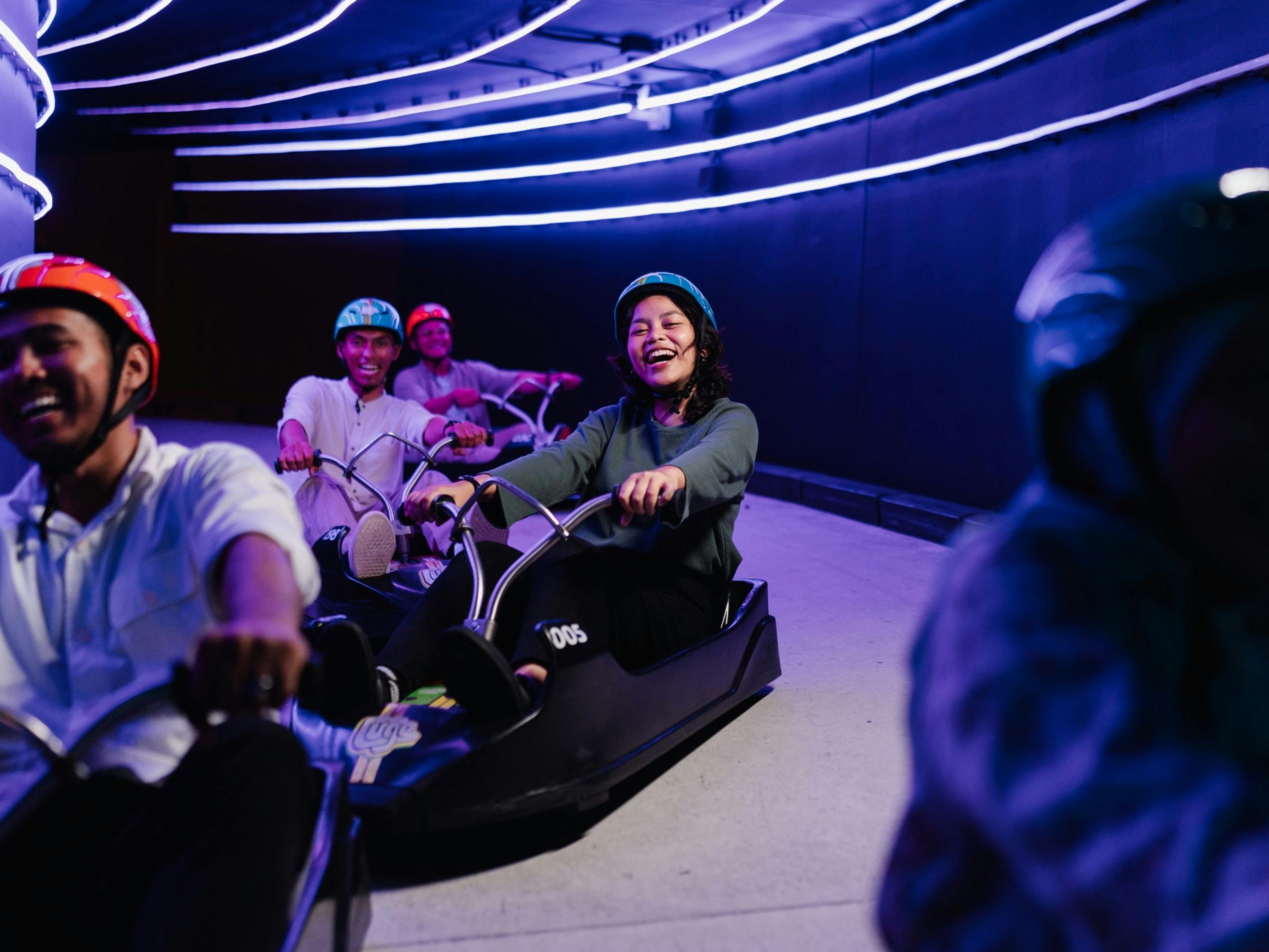 People riding go-karts indoors under illuminated curved light lines, smiling and enjoying the activity.