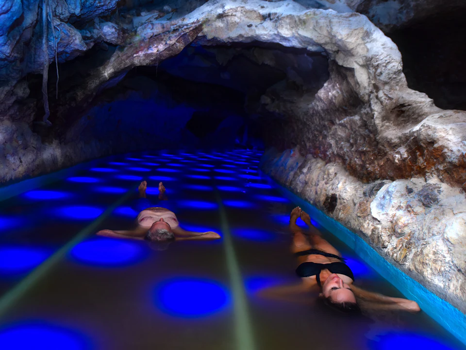 Two individuals relaxing in a shallow, illuminated cave pool with blue circular light patterns on the water surface.