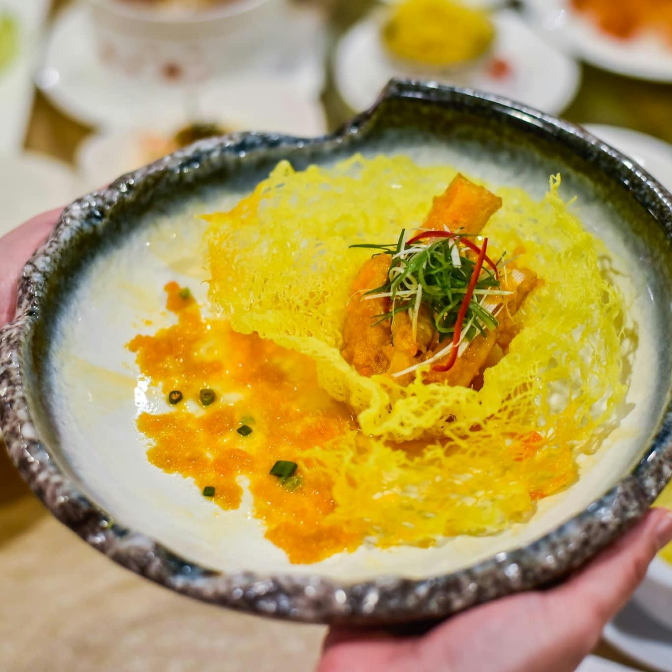 A plated dish featuring crispy yellow garnish, fried item, and sauce, presented in a textured bowl being held by hands.
