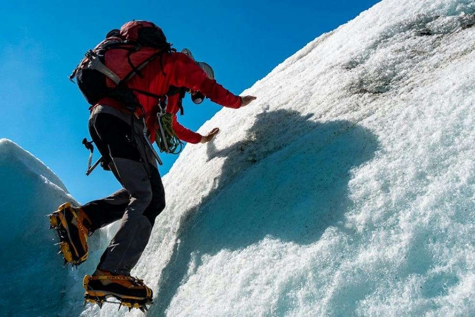 A climber wearing a red jacket and crampons ascends a steep icy slope under a clear blue sky.