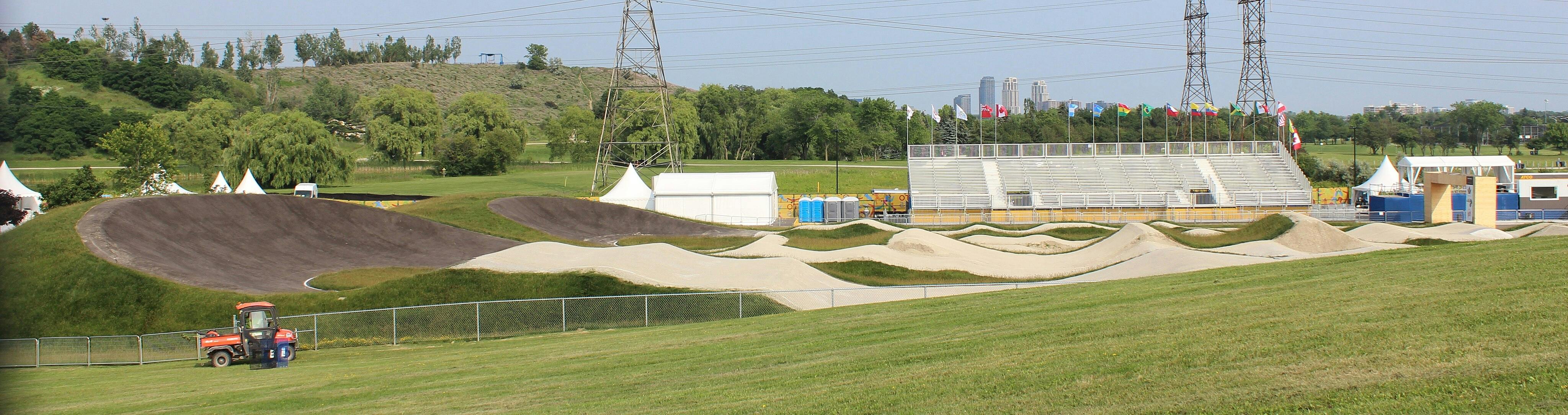 A BMX track with undulating terrain, a grandstand with international flags, tents, and portable toilets in a park setting.