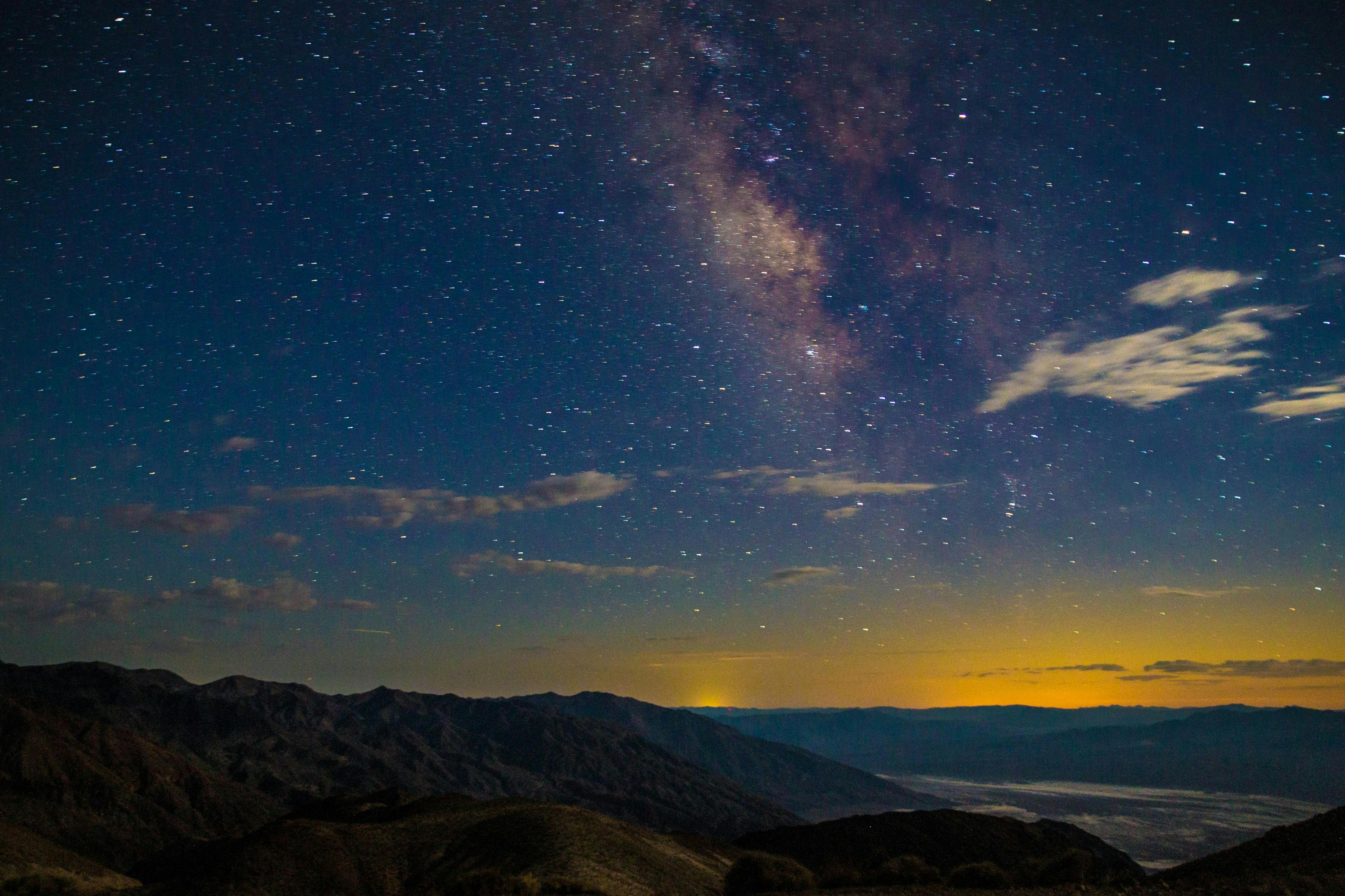 A starry night sky with the Milky Way visible, above a mountainous landscape and a faintly glowing horizon.