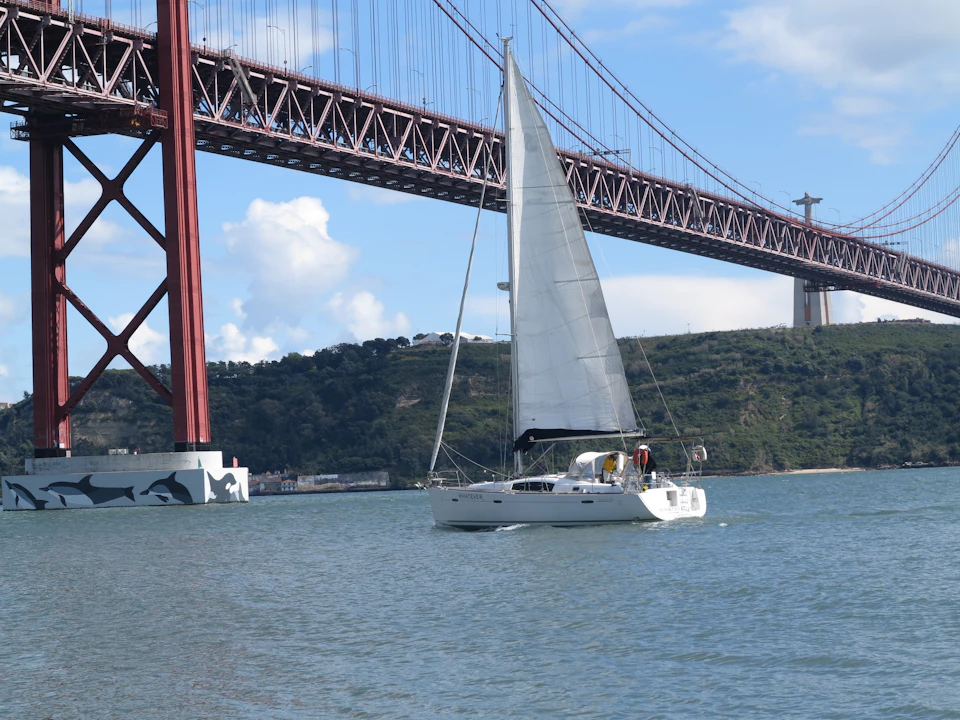 A sailboat with white sails glides on water beneath a large red suspension bridge on a clear day. Hills and clouds are in the background.