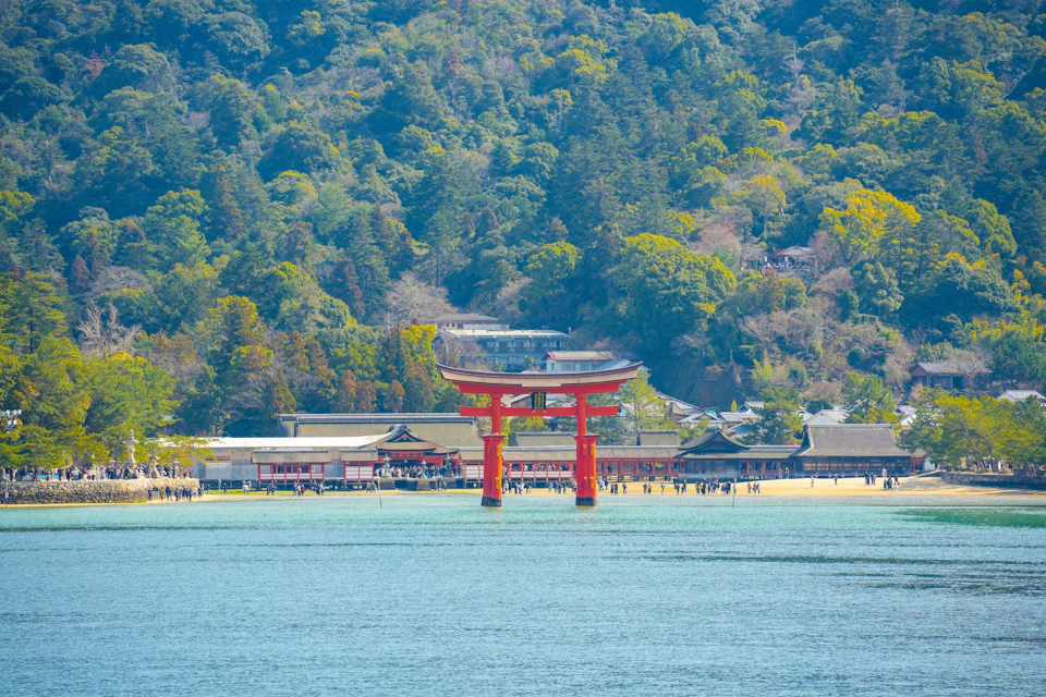 A red torii gate stands in shallow water with a traditional Japanese shrine and a forested hillside in the background.