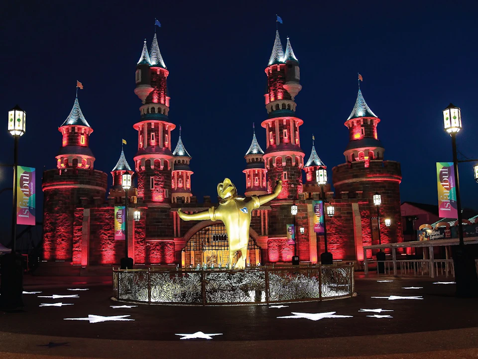 Illuminated castle with turrets at night, featuring a central golden statue of a figure with raised arms in front. White star patterns on the ground.