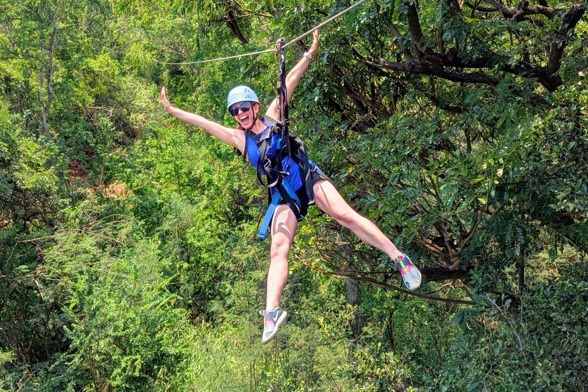 A person in a blue outfit and helmet ziplining through a lush green forest, smiling with arms outstretched.