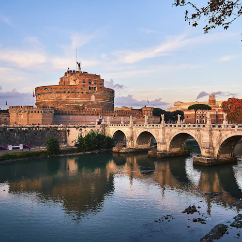 Historisk stenbro med valv sträcker sig över en flod och leder till ett stort cylindriskt slott. Höstträd och byggnader i bakgrunden.