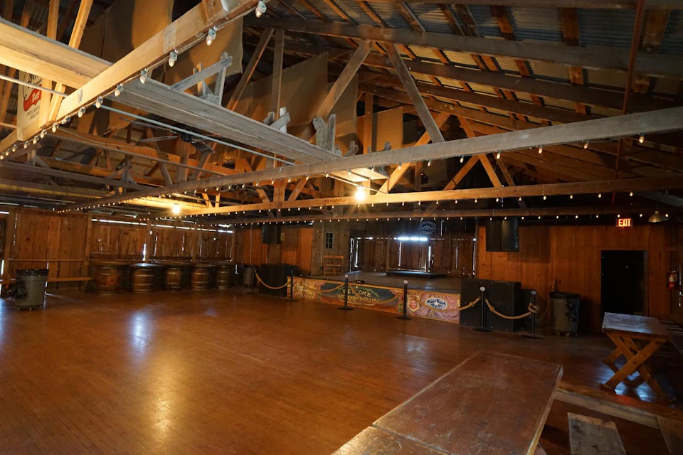 A rustic, empty wooden hall with a raised stage, string lights, wooden barrels, and large wooden beams supporting the ceiling.