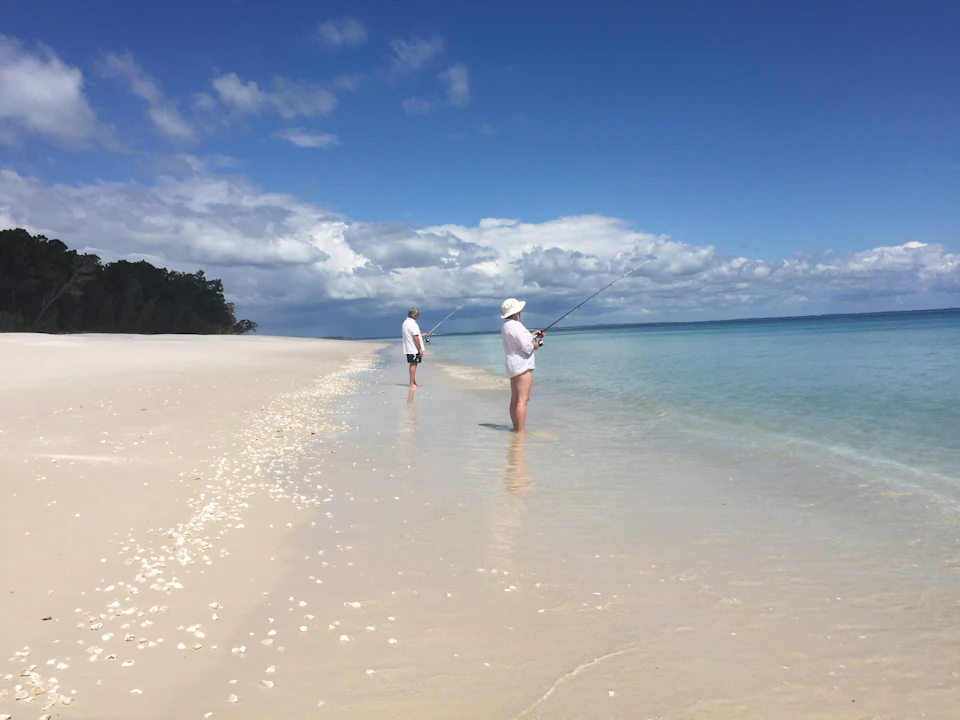 Two people in white hats and clothing fish while standing in shallow, clear water on a wide sandy beach under a partly cloudy sky.