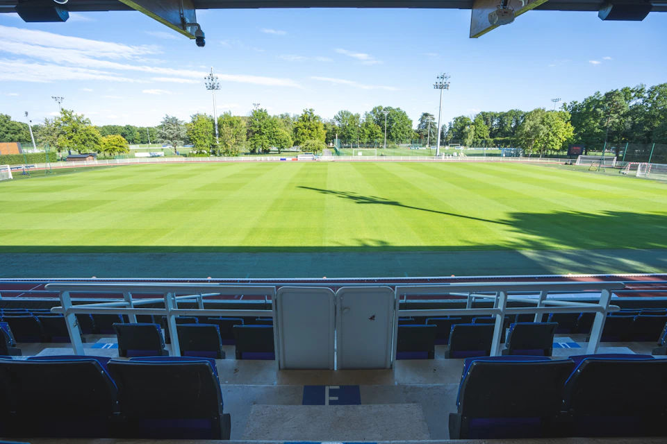 A sports field viewed from the seating area with green grass, surrounding trees, and a clear blue sky.