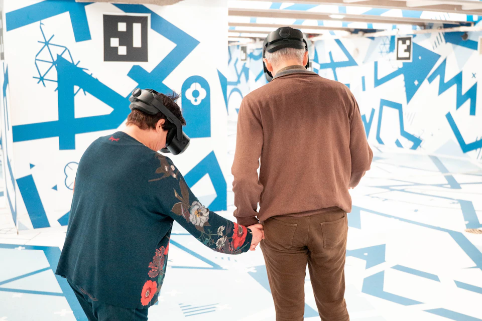 Two people wearing VR headsets interact in a room with abstract blue and white patterns on the walls and floor.